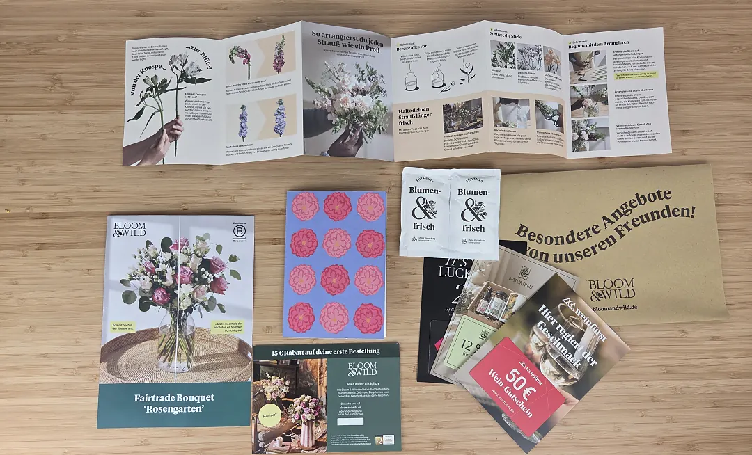 Alles aus dem Bloom & Wild Karton auf einem Tisch: Anleitung zum Arrangieren, Strauß-Karte, Grußkarte, zwei Tütchen Blumennahrung und Gutscheine
