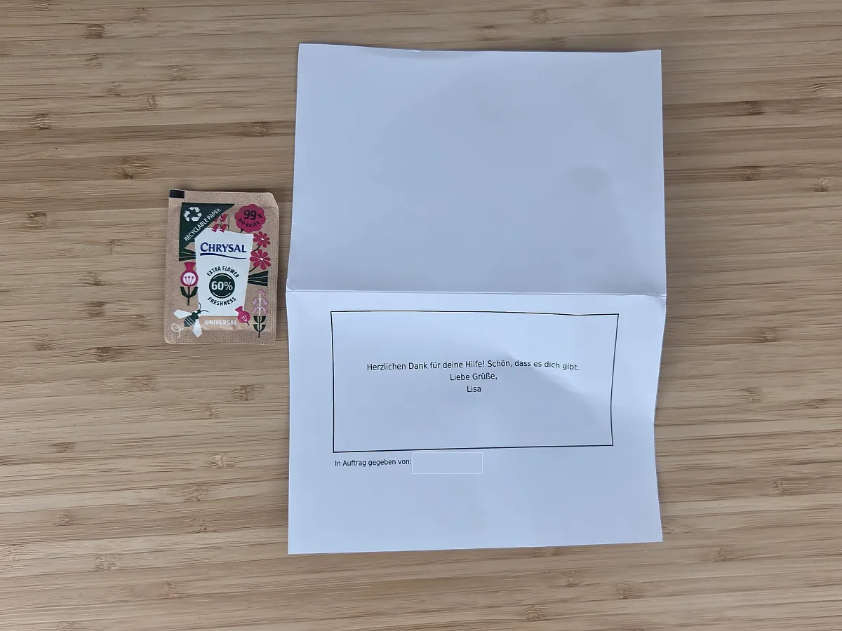Bloomy Days Grußkarte als bedrucktes Papier mit dem Hinweis In Auftrag gegeben von, der Name ist abgedeckt, daneben ein Tütchen Blumennahrung